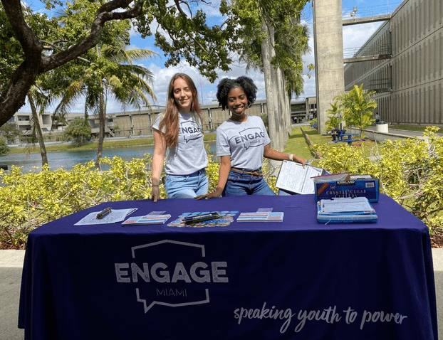 Two young people are shown tabling for Engage Miami to encourage their peers to register to vote so that their voices can be heard. 