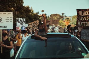 A young Black girl in a COVID mask raises her fist, surrounded by people carrying signs that read "BLACK LIVES MATTER"