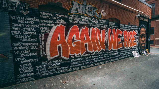 A brick wall is decorated by graffiti reading, "Again we rise" surrounded by the names of over 100 people who were killed due to police brutality.
