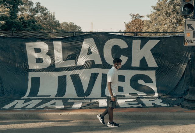 A masked individual walks in front of a Black Lives Matter banner. 