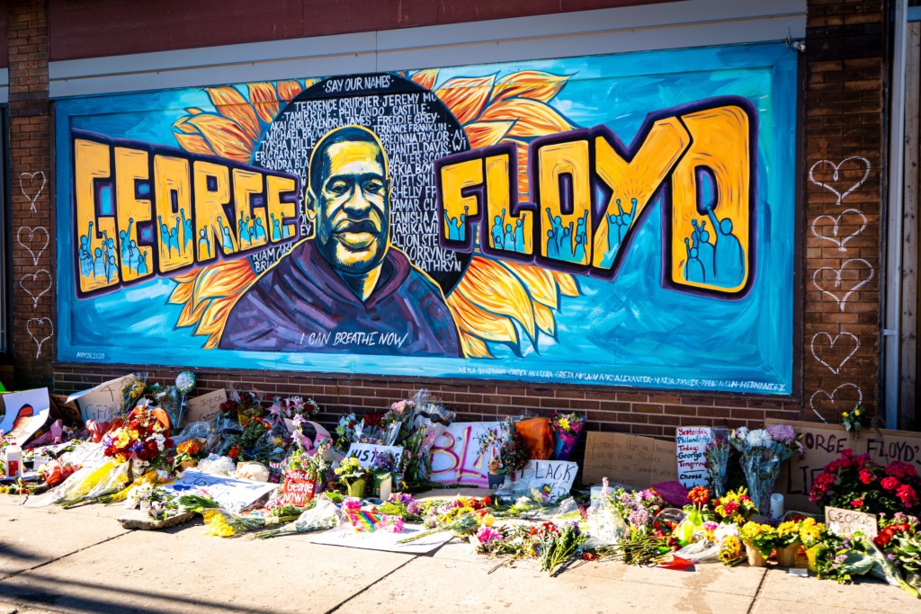 Photo of a George Floyd mural with flowers and signs on the sidewalk.