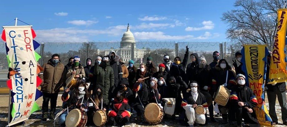 A group takes a picture in from of the US Capitol building with drums and signs. One sign reads "Citizenship For All"