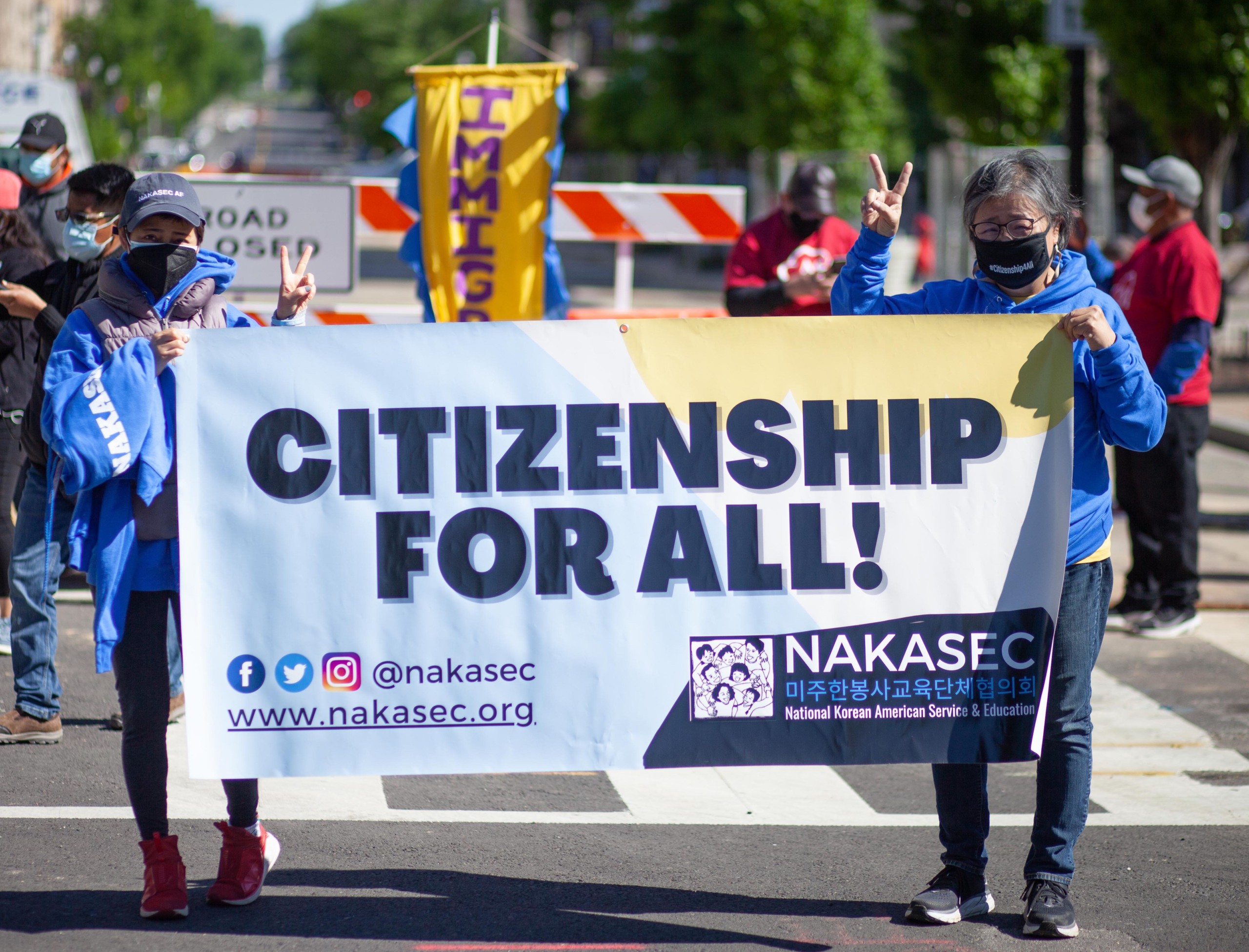 Two individuals at a rally hold up a banner that says "Citizenship for All!"