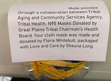 A label is shown on an individually-wrapped COVID mask that reads, "Made possible through a collaboration between Tribal Aging and Community Services Agency, Tibal Health, N95 Masks Donated by Great Plains Tribal Charman's HEalth Board. Your vloth mask was made and donated by Floris Whitebull, packaged with Love and Care by Shauna Long."