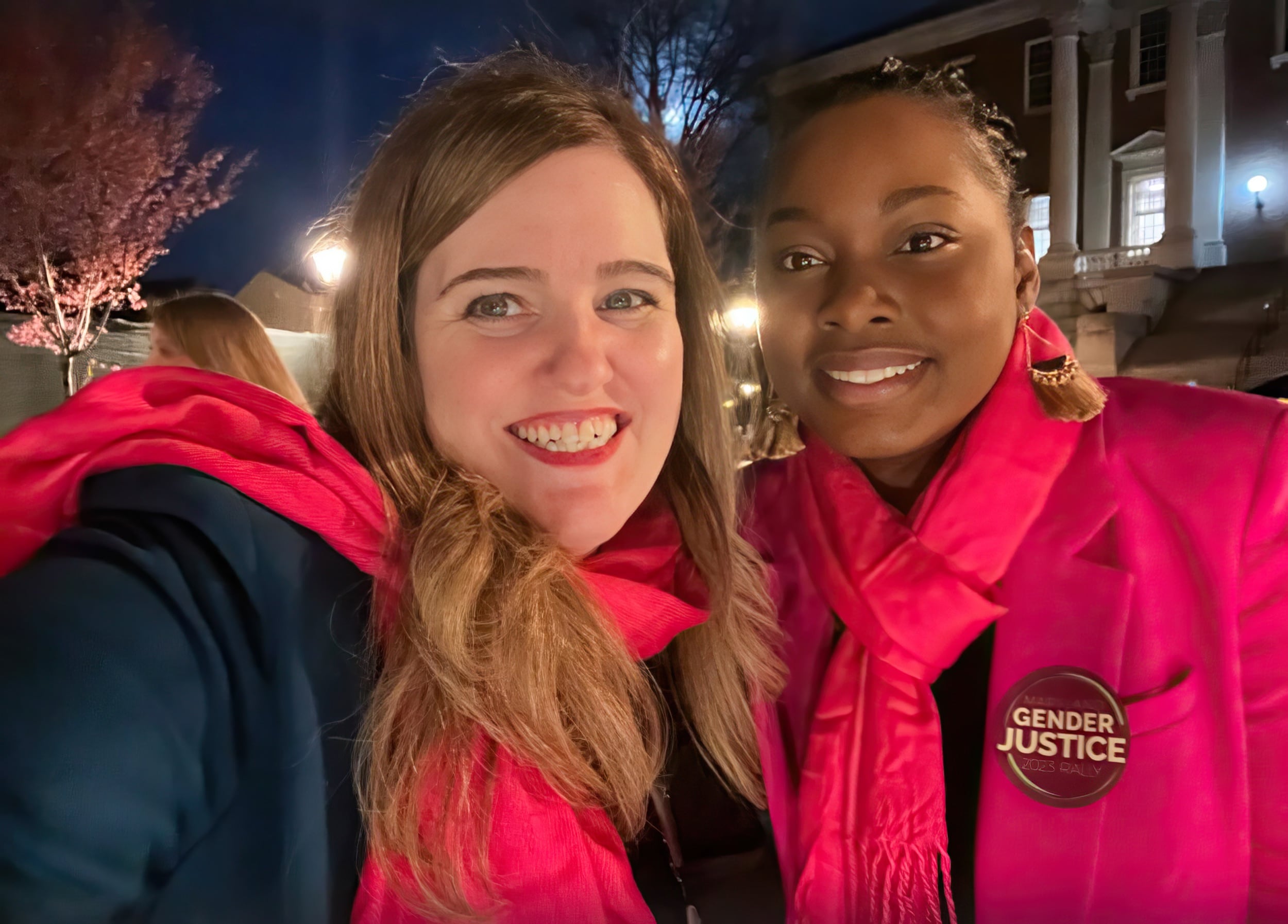 Jakeya Johnson and a second woman both wearing bright pink and smiling. Jakeya is wearing a pin on her blazer that reads ‘gender justice.’