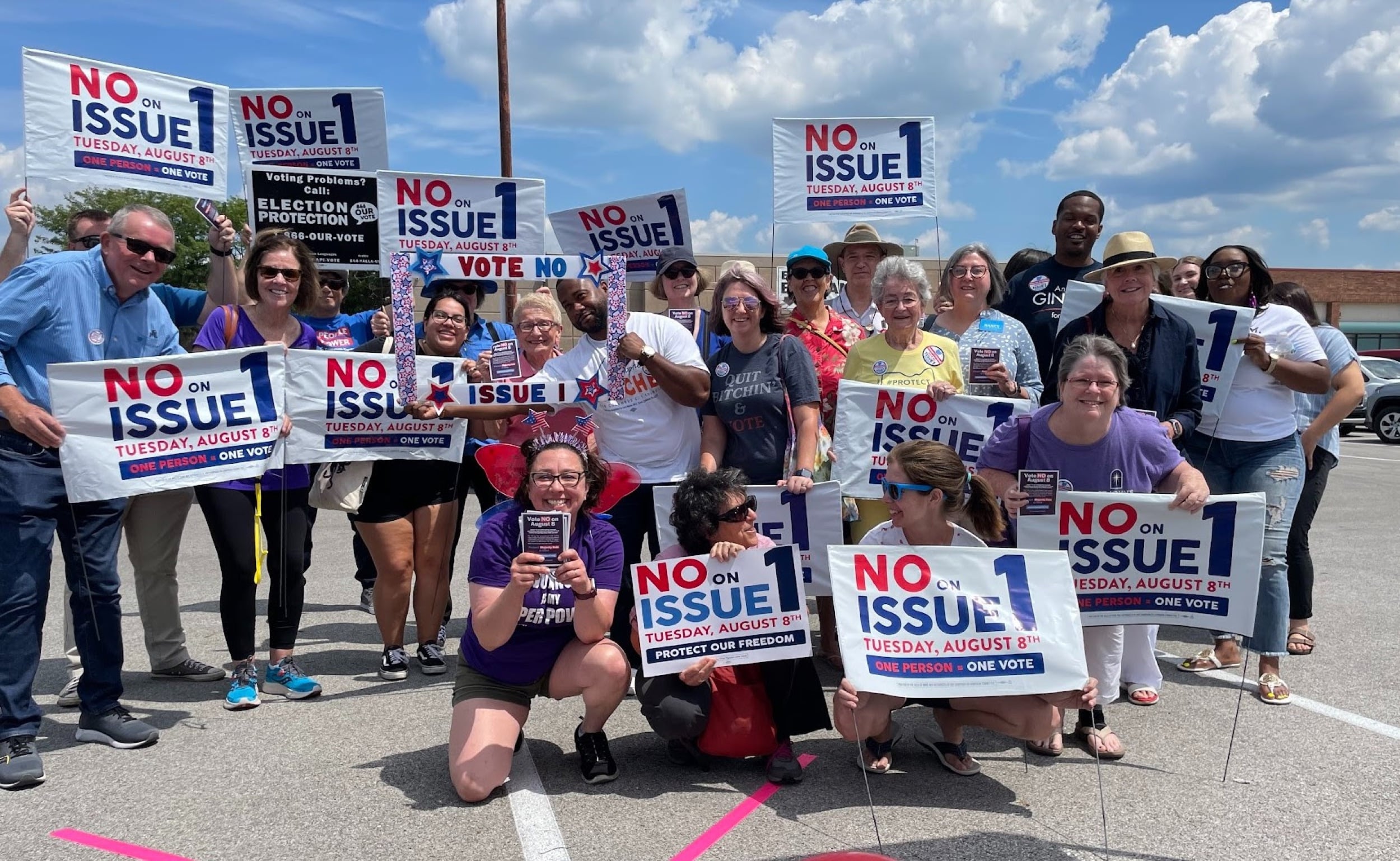 A large group of multi-racial and multigenerational community members posing together in a parking lot, holding signs that read ‘No on Issue #1.’