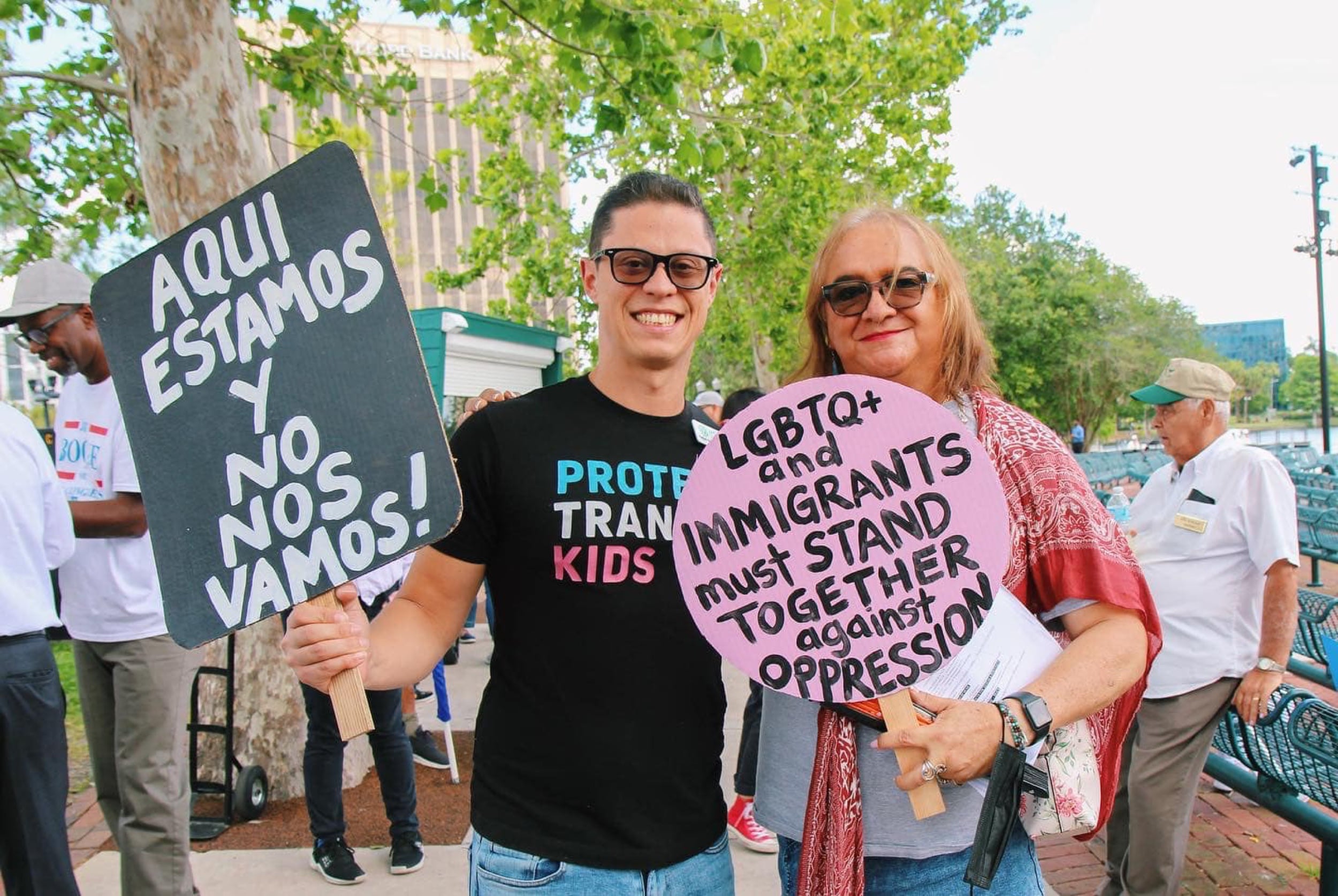 Two protesters holding signs that read 'Aqui estamos y no nos vamos!' and 'LGBTQ+ and immigrants must stand together against oppression.' 