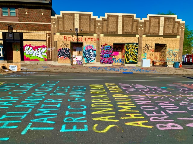 A graffiti-lined urban scene is punctuated by the names of those killed by police brutality: Jamar Clark, Philando Castile, Tamir Rice, Eric Garner, Sandra Bland, Ahmaud Arbery, Breonna Taylor, Alton Sterling, and others. 
