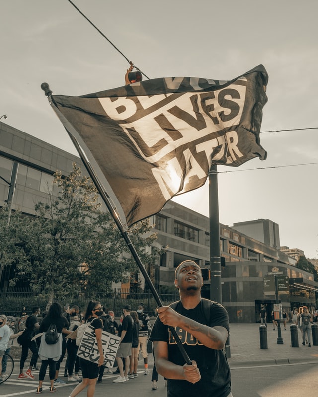 A man proudly waves a "BLACK LIVES MATTER" flag high above his head during a protest decrying violence against Black Americans amidst ongoing police brutality.