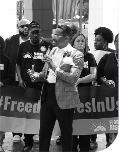Black man in a suit speaking into a microphone in front of a crowd wearing Florida Rising shirts and holding placards and banners.