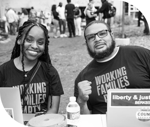 Black woman and man wearing glasses seated at a table with laptops, both wearing Working Families Party shirts.