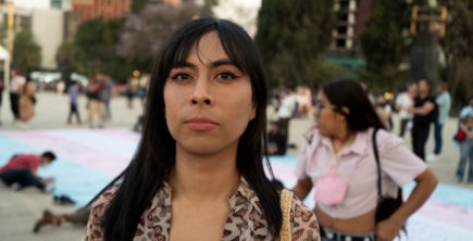 A Latine woman standing proudly in front of a large trans flag banner.