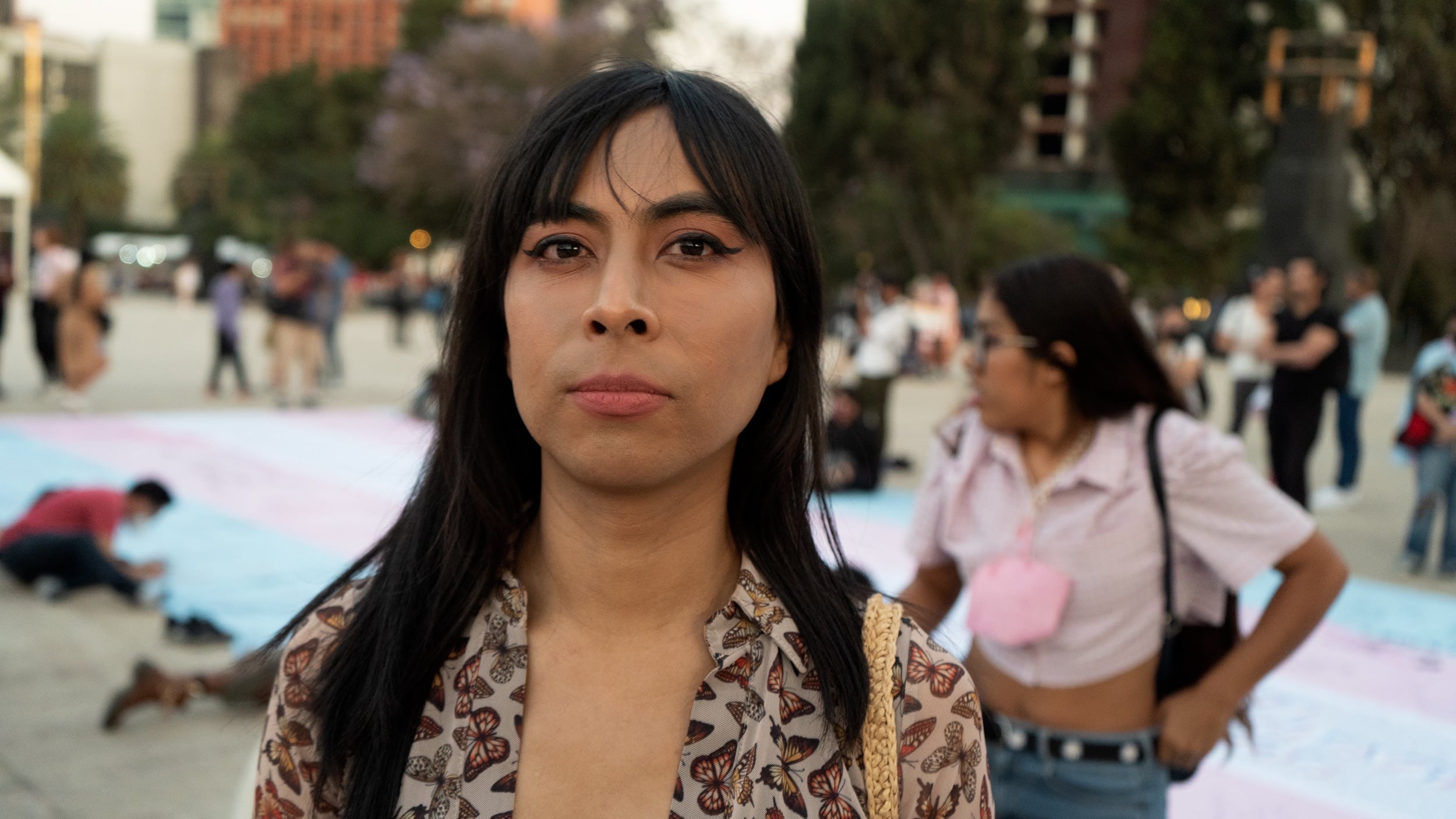 A Latine woman standing proudly in front of a large trans flag banner.