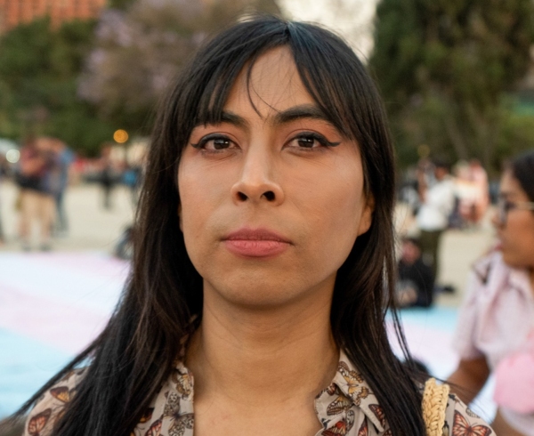 A Latine woman standing proudly in front of a large trans flag banner.