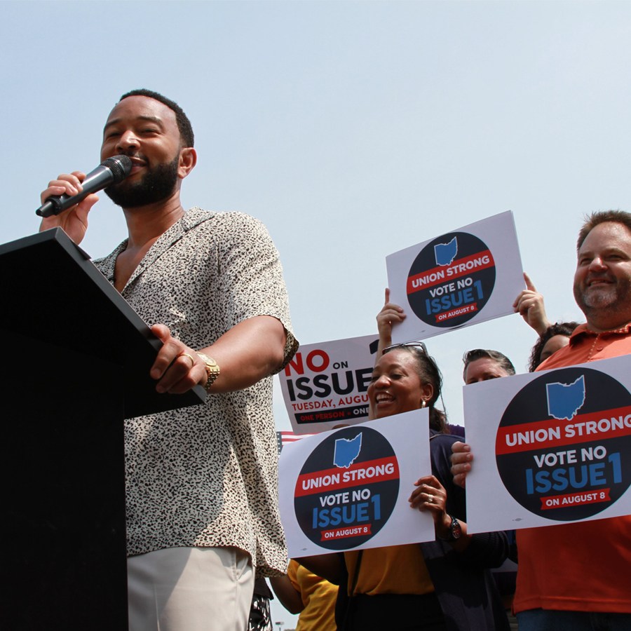 DC-Aug-2023-square John Legend standing at a podium holding a microphone with a multi-racial group of people behind him holding up signs that read ‘Union Strong. Vote Issue #1.’