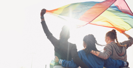 Three people holding a rainbow Pride flag above their heads. Wind is blowing the flag.