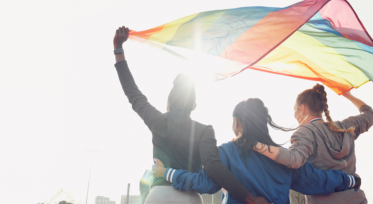 Three people holding a rainbow Pride flag above their heads. Wind is blowing the flag.