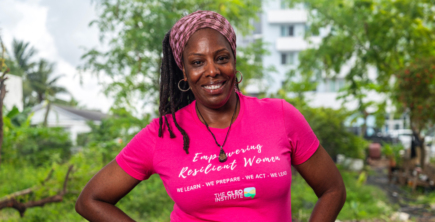 Black woman smiling, standing outside against a green tree with her arms on her hips. She's wearing a bright pink tshirt that says Empowering Resilient Women Cleo Institute.