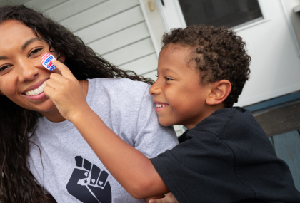 A Black woman wearing a grey T-shirt with a Black power fist and a young Black child is pressing a 'I voted' sticker onto her cheek as she smiles