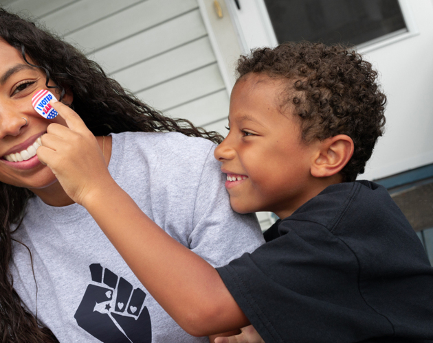 A Black woman wearing a grey T-shirt with a Black power fist and a young Black child is pressing a 'I voted' sticker onto her cheek as she smiles