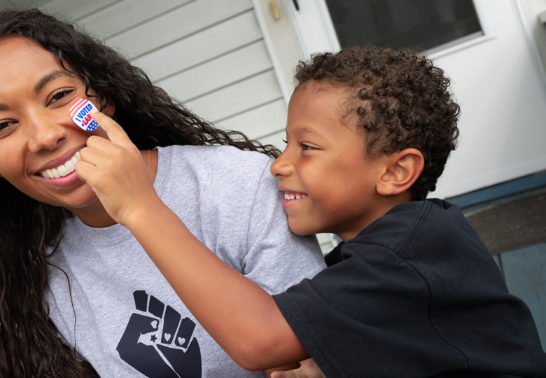 A Black woman wearing a grey T-shirt with a Black power fist and a young Black child is pressing a 'I voted' sticker onto her cheek as she smiles