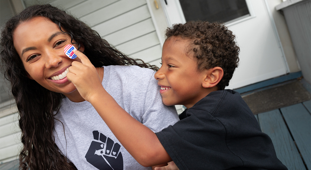 A Black woman wearing a grey T-shirt with a Black power fist and a young Black child is pressing a 'I voted' sticker onto her cheek as she smiles