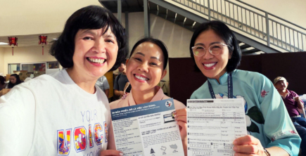 Three smiling community members in Houston hold Vietnamese-language voter registration forms and information about the November 2026 election.